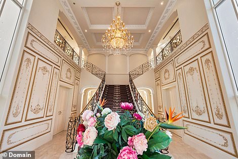 Through the arched double doors decorated with swirling gold iron, there is a grand staircase with an ornate balustrade, decorative wainscoating and a sparkling chandelier